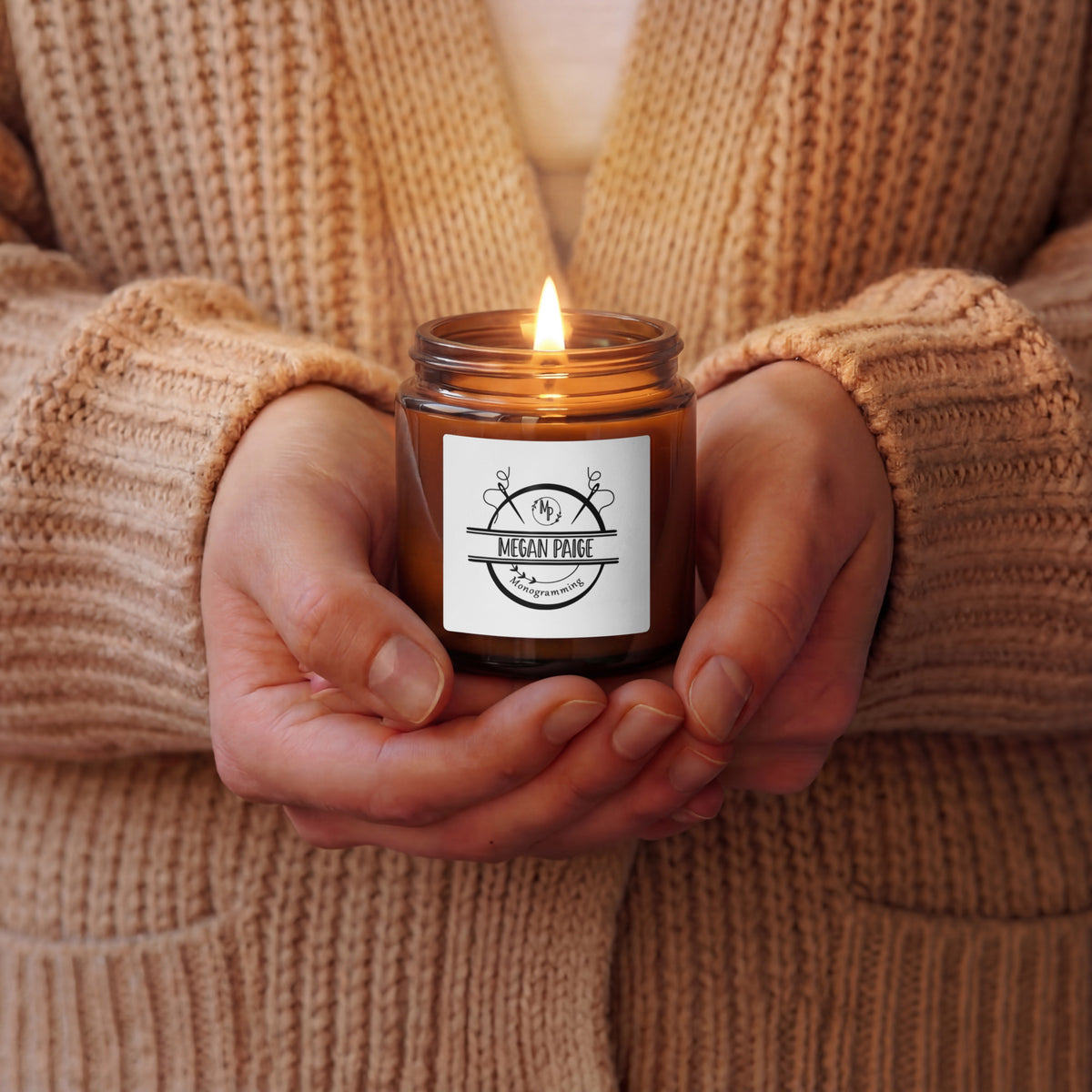 Person holding a lit candle in a brown jar with a label, wearing a beige sweater.