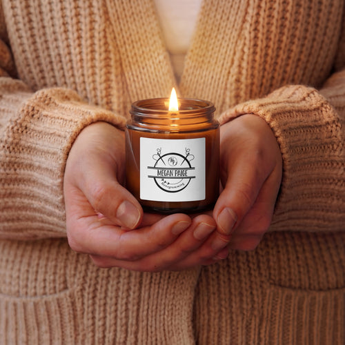 Person holding a lit candle in a brown jar with a label, wearing a beige sweater.