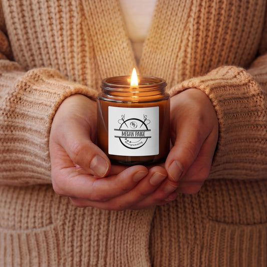 Person holding a lit candle in a brown jar with a label, wearing a beige sweater.